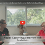 Two individuals are sitting at a table with a centerpiece in a room with a large window Text overlay reads, "Flagler County Buzz Interview with Cornelia Manfre on her Bid for Palm Coast Mayor " A red play button indicates a video